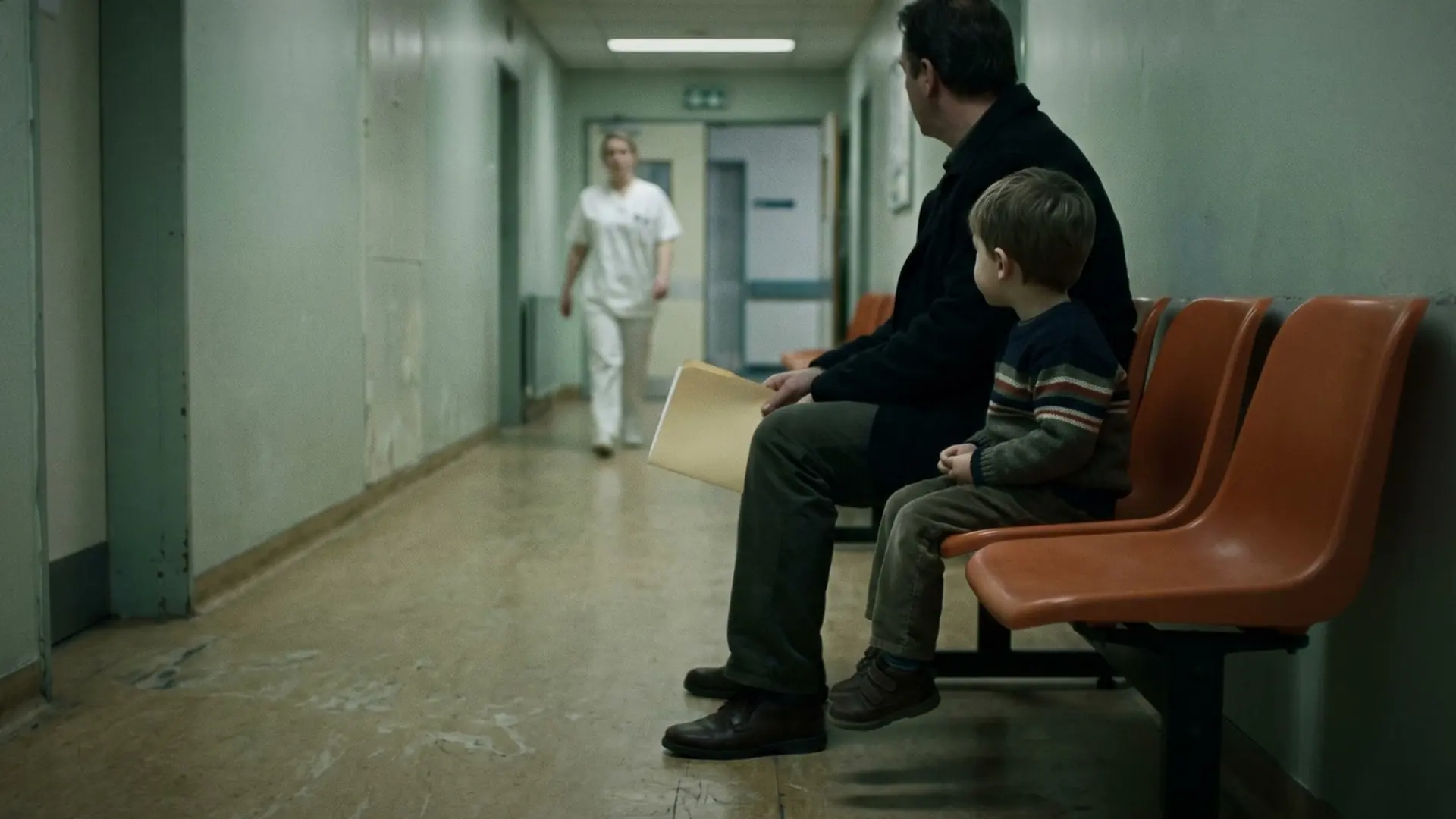 A man and a small boy waiting on orange chairs in a pale green clinic corridor.