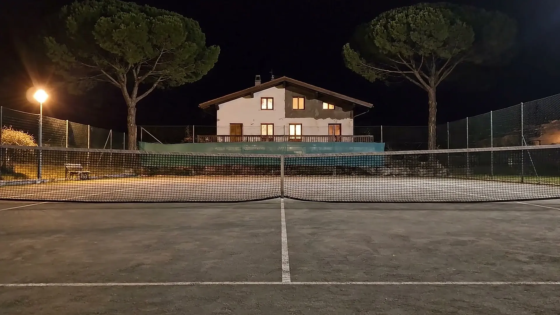 An empty tennis court at night, with a house and two umbrella pines behind it.