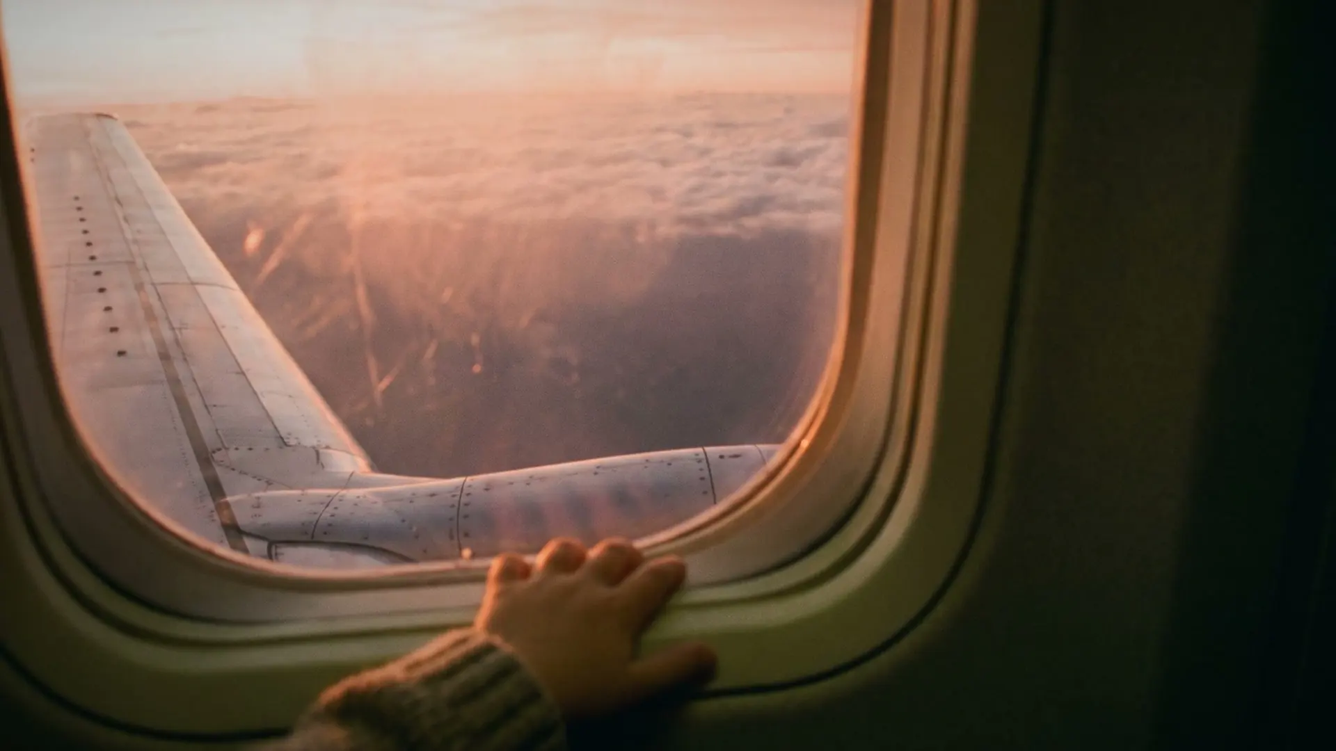A hand on an aeroplane window, with the wing and pink clouds outside.