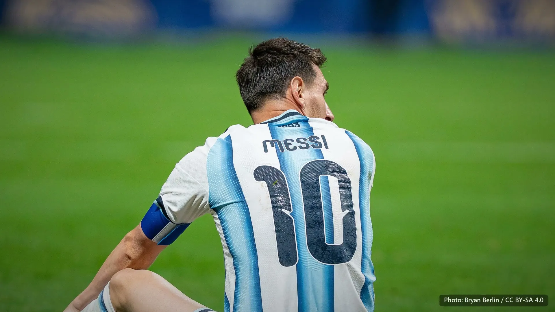 Messi seated on the grass, seen from behind and turning his head, in Argentina's 10 shirt.